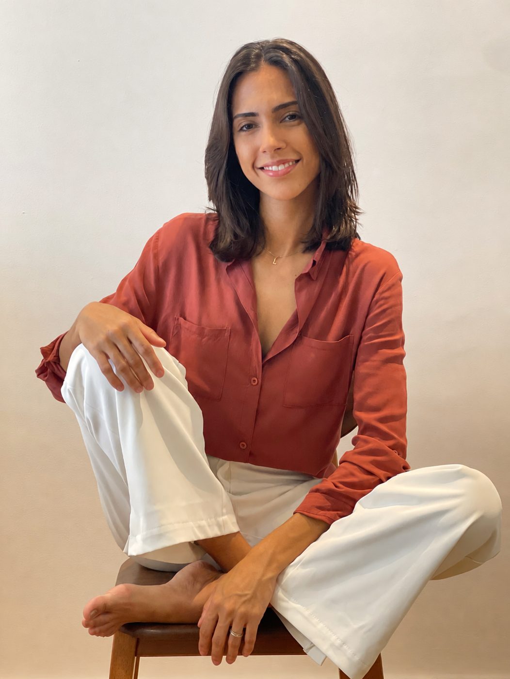 Lya Morena sorrindo, sentada com camisa terracota e calça branca diante de uma parede clara.
